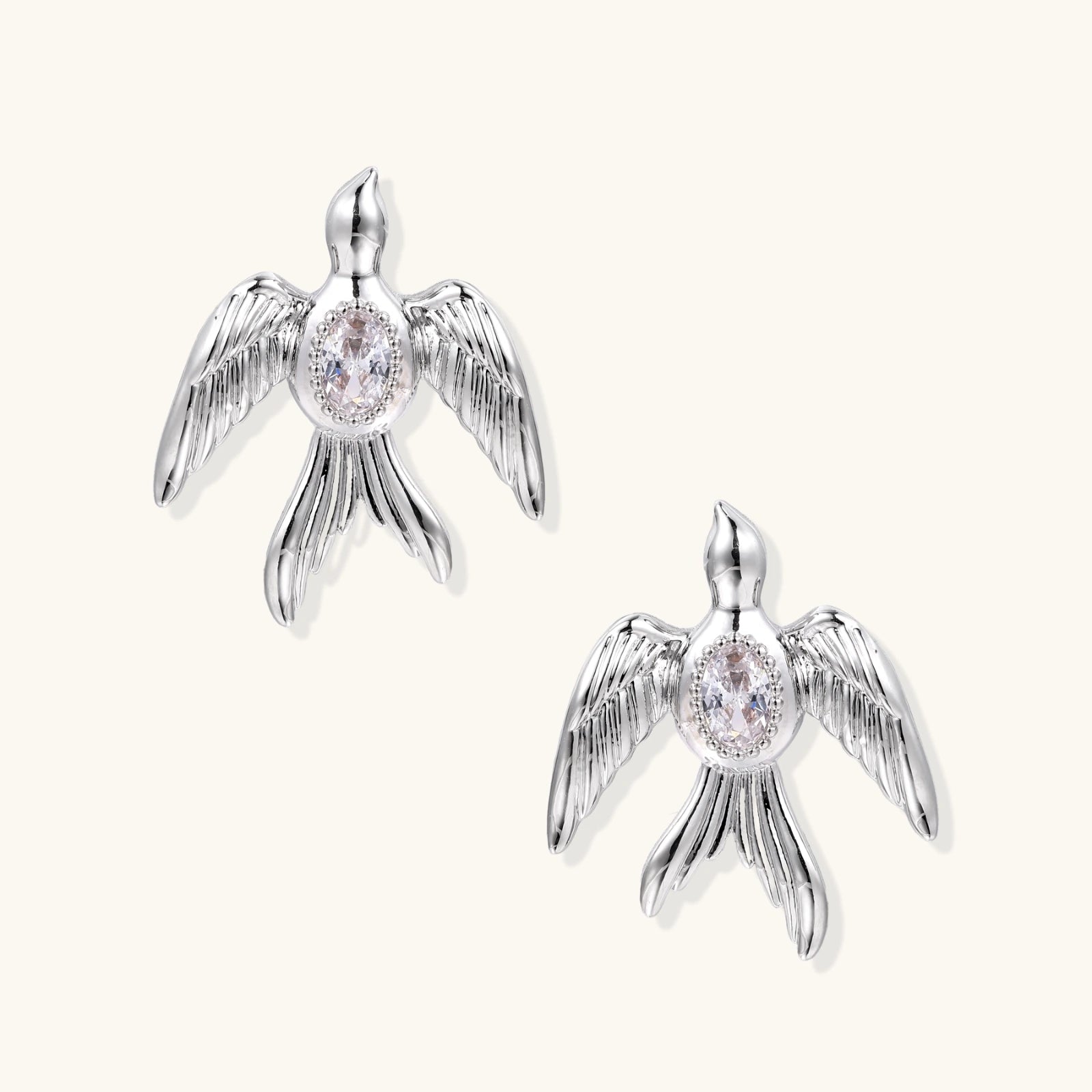 White gold-plated bird earrings with spread wings and oval zirconia center on cream background