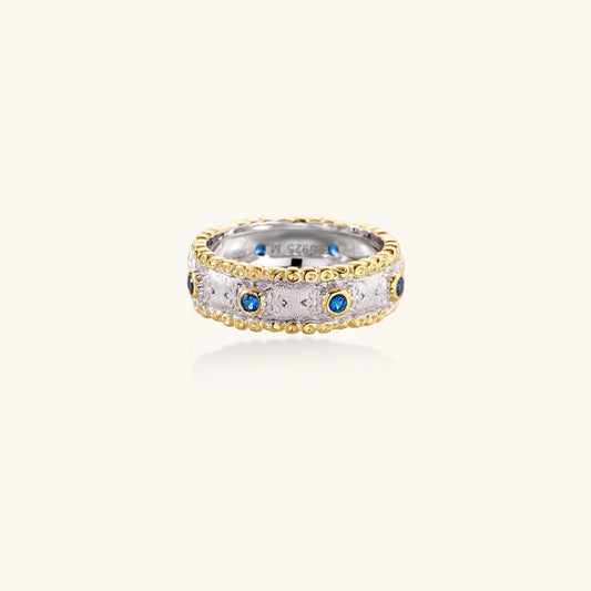 Two-tone ring with brushed silver center, gold beaded edges, and blue zirconia stones on white background.
